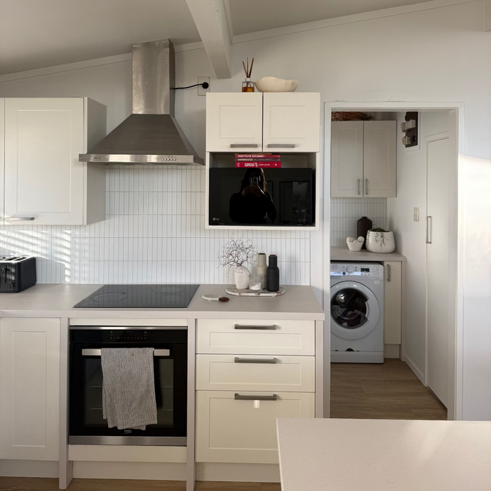 A modern kitchen interior with white cabinets, a stainless steel range hood, built-in black appliances, a tiled backsplash, and a washing machine visible in an adjacent room.