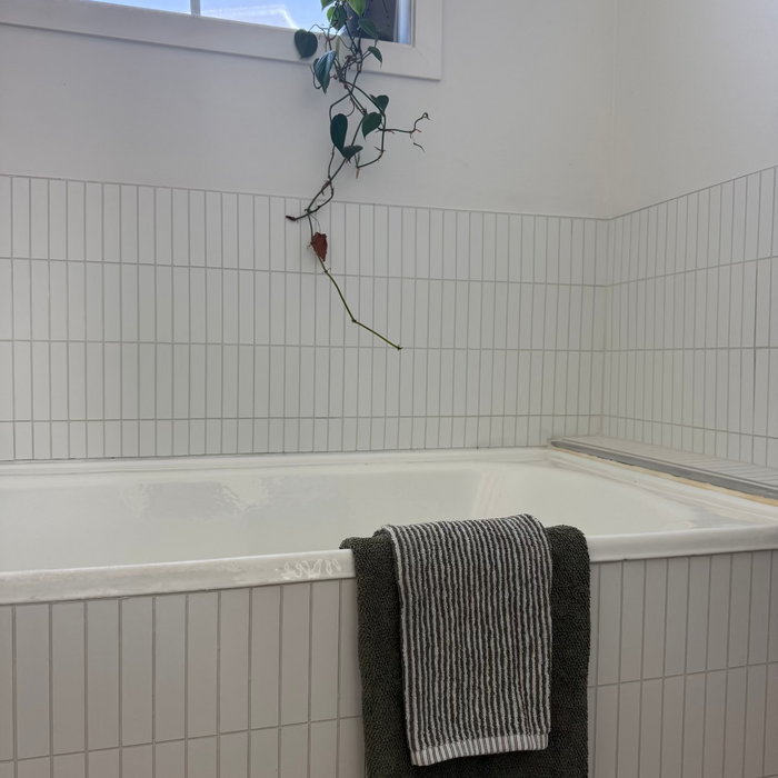 A white tiled bathroom with a built-in bathtub, a dark green bath towel and a striped gray-and-white towel draped over the tub edge, and a small trailing plant hanging near the window.