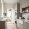 A modern laundry room with light grey cabinets, a white farmhouse sink, chrome faucet, stacked towels, and built-in washing machines. An open doorway at the back leads to a small patio area with potted plants.