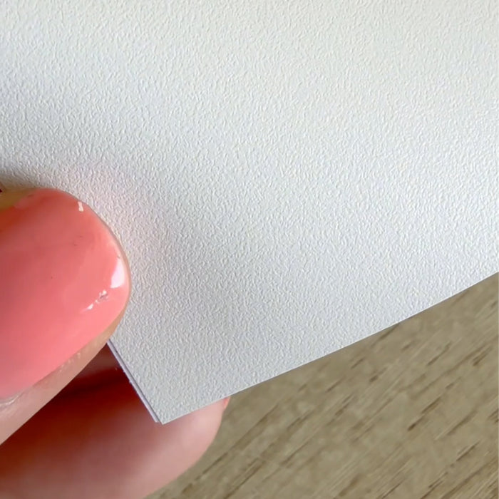 A close-up of textured apricot grey adhesive vinyl held by a hand with pink nail polish, with a light wood surface visible in the background.