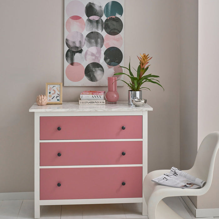 A white wooden dresser with three pink drawer fronts and small black knobs, styled in a room setting with a plant, books, and a framed print, next to a modern white chair.