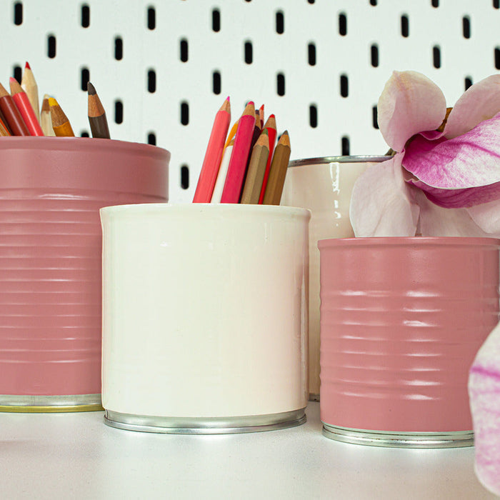 Three ribbed metal can-style containers in dusty pink and off-white, arranged on a white surface against a white pegboard background. The pink containers hold colored pencils, and a pink orchid flower is placed in the right container.