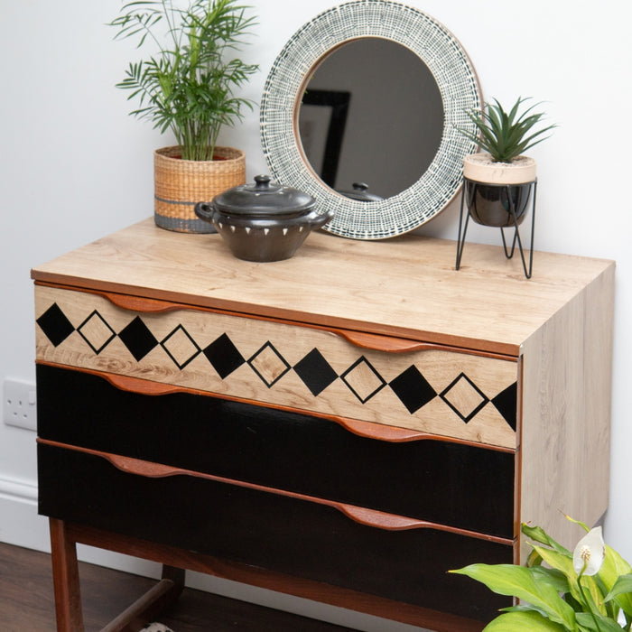 A wooden dresser with flower pots and a round mirror and black glossy vinyl wrapped drawer fronts.