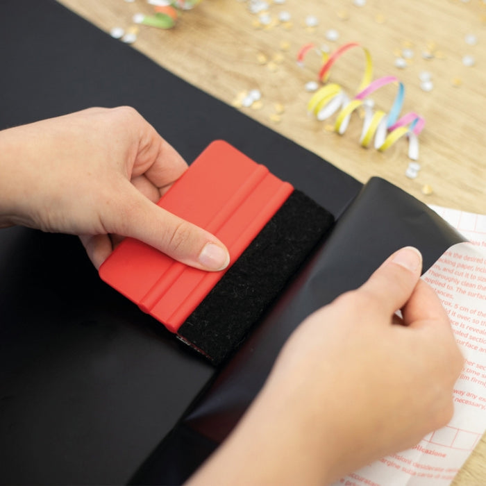 A hand holding a red squeegee installing black matte adhesive vinyl onto a panel.