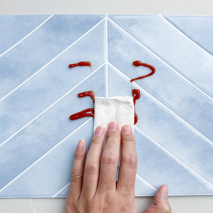 Hand using a paper towel to clean up ketchup spills on a blue chevron tile.