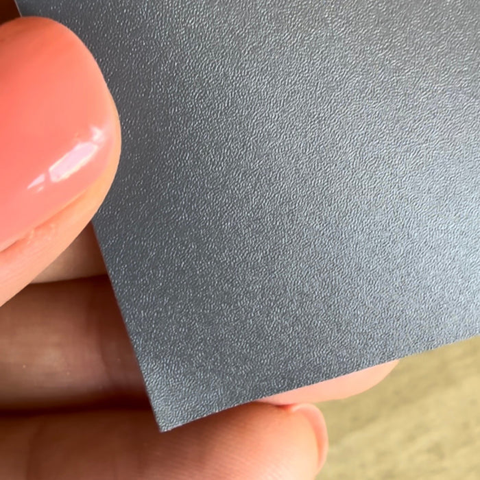 A close-up of a hand holding a deep grey textured adhesive vinyl, showing the surface detail and edge against a blurred beige background.