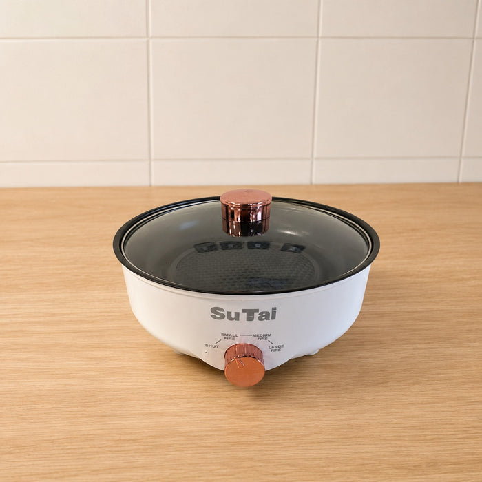 A Electric Frying Pan with a clear glass lid and copper-colored knobs, sitting on a light wood countertop against a white tiled wall.