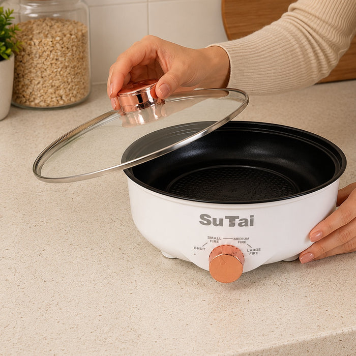 A white SuTai eElectric Frying Pan with a black non-stick interior, a clear glass lid with a rose gold handle, and a rose gold rotary dial, shown on a kitchen countertop with a person's hands interacting with it.