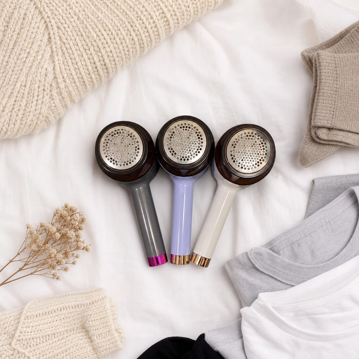 USB Rechargeable Fabric Lint Remover with different color handles on a white surface with clothing and yarn.