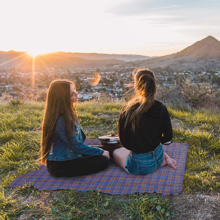 Two people sitting on a Foldable Waterproof Picnic Mat overlooking a town with mountains at sunset.
