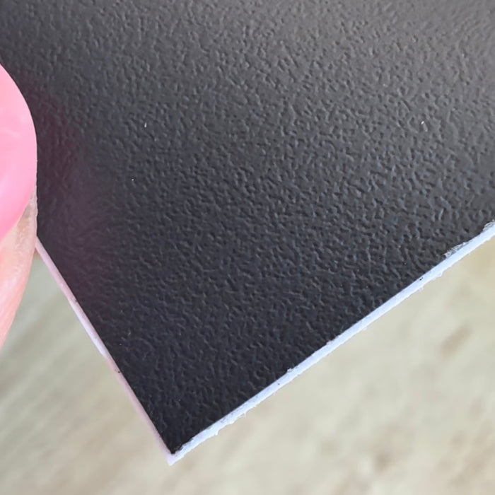 A close-up of meteorite grey textured adhesive vinyl, showing its pebbled surface and white backing edge against a light wood background.