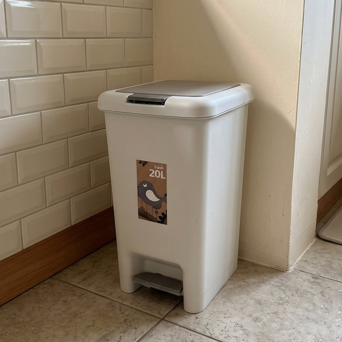 A 20L plastic waste bin with a foot pedal, placed in a kitchen corner against a cream subway tile wall and tiled floor.