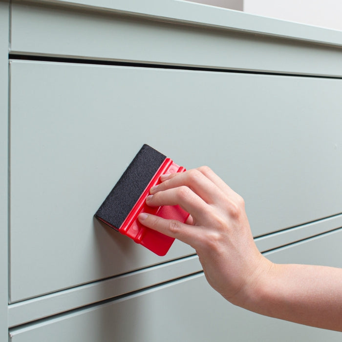 A hand holding a red squeegee over a sage green vinyl wrapped dresser.