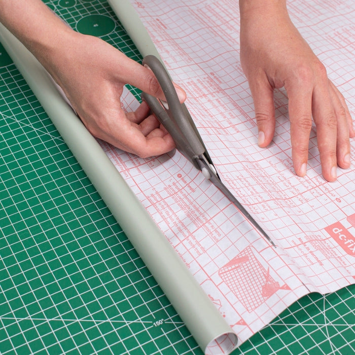 A hand cutting off a piece of sage green adhesive vinyl using the gridded backing paper as a guide.
