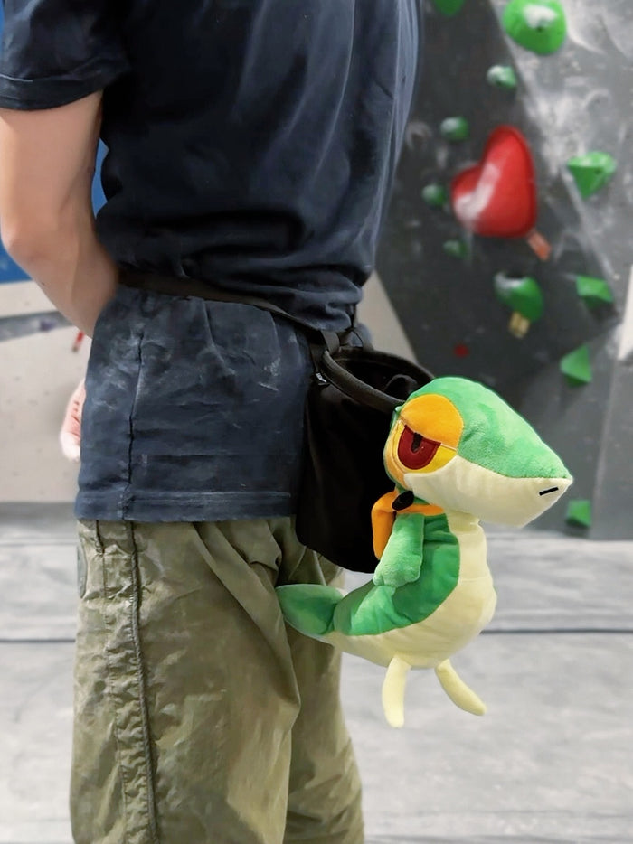 A cute, handmade plush climbing chalk bag designed to look like the Snivy, featuring a sleek green and cream body with yellow leaf-like accents and large, confident eyes. Designed with high functionality by a climber, it includes a wide black rim opening at the top for easy chalk access. A great climbing gift for trainers who want to catch them all.