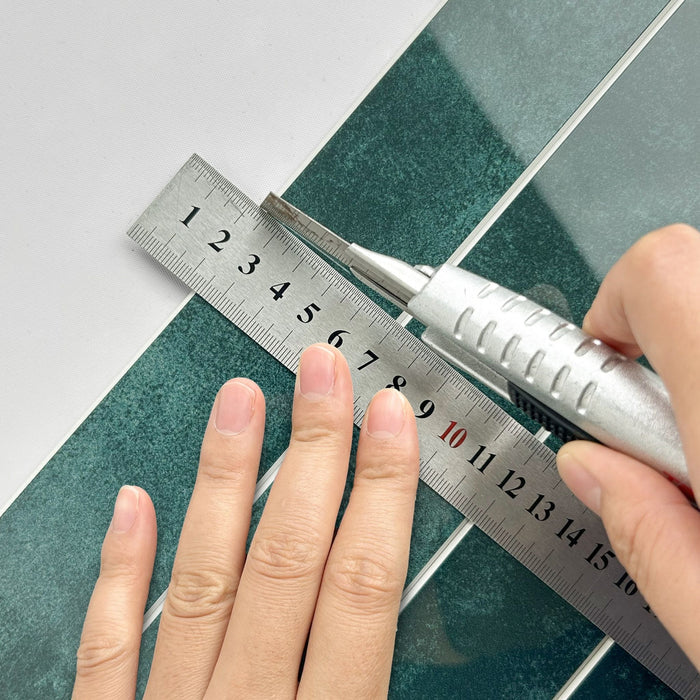 A close-up of a hand using a silver metal ruler and utility knife to measure and cut a dark green composite tile, on a white background.