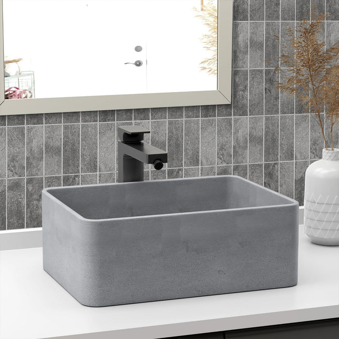 A rectangular grey concrete bathroom vessel sink with a matte black faucet, set on a white countertop against a grey tiled backsplash.