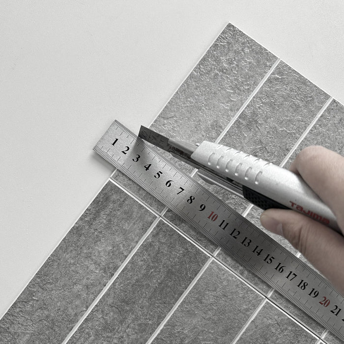 A close-up of a hand using a utility knife to cut a sheet of grey textured tile along a metal ruler.