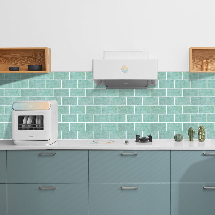 A modern kitchen scene with a white countertop, mint green subway tile backsplash, pale blue cabinets, a white range hood, and a white countertop appliance on the left.