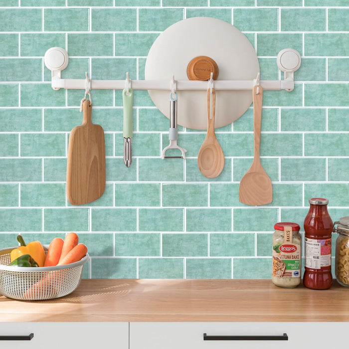 A white wall-mounted kitchen utensil rack with suction cups is shown installed on a mint green subway tile backsplash above a wooden countertop. Hanging from the rack are wooden cutting and serving utensils, including a paddle, spoon, spatula, and peeler, along with a round white holder.