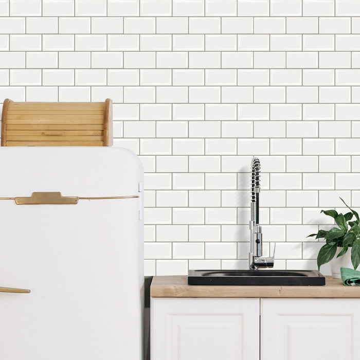 White subway tile backsplash installed behind a kitchen sink with a chrome faucet, light wood countertop, white cabinets, and part of a white refrigerator with gold handles.