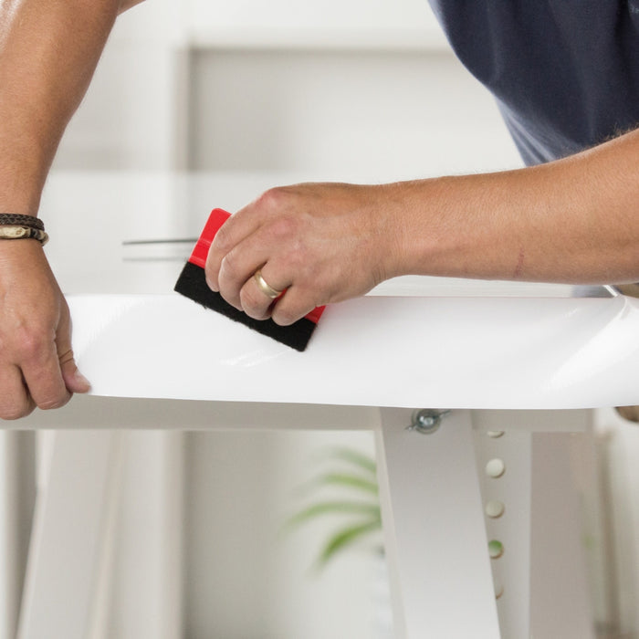 A person's hands using a squeegee to apply white glossy adhesive vinyl to a furniture surface.